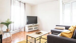 En stue med et fladskærms-tv på et hvidt stativ, en grå sofa med puder, et lille træsofabord og en potteplante.