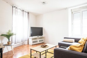 Ein Wohnzimmer mit einem Flachbildfernseher auf einem weißen Schrank, einem grauen Sofa mit Kissen, einem kleinen Holztisch und einer Topfpflanze.