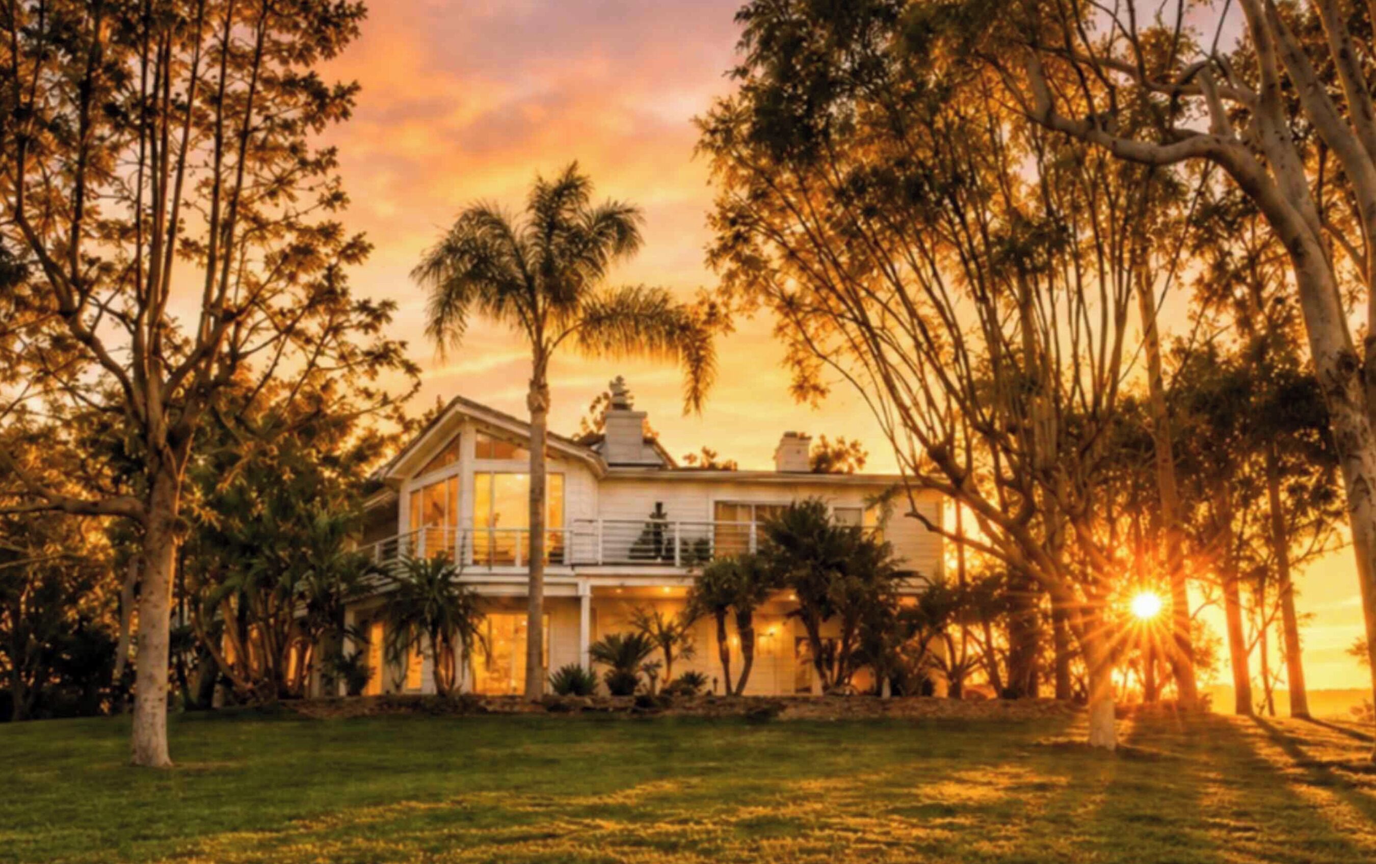 A two-story house with a balcony, surrounded by trees and a lawn, during sunset.