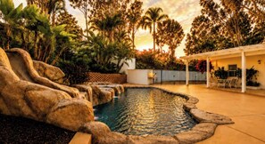 En pool med en naturlig stenformation som designelement, omgiven av frodig grönska och en vit pergola.