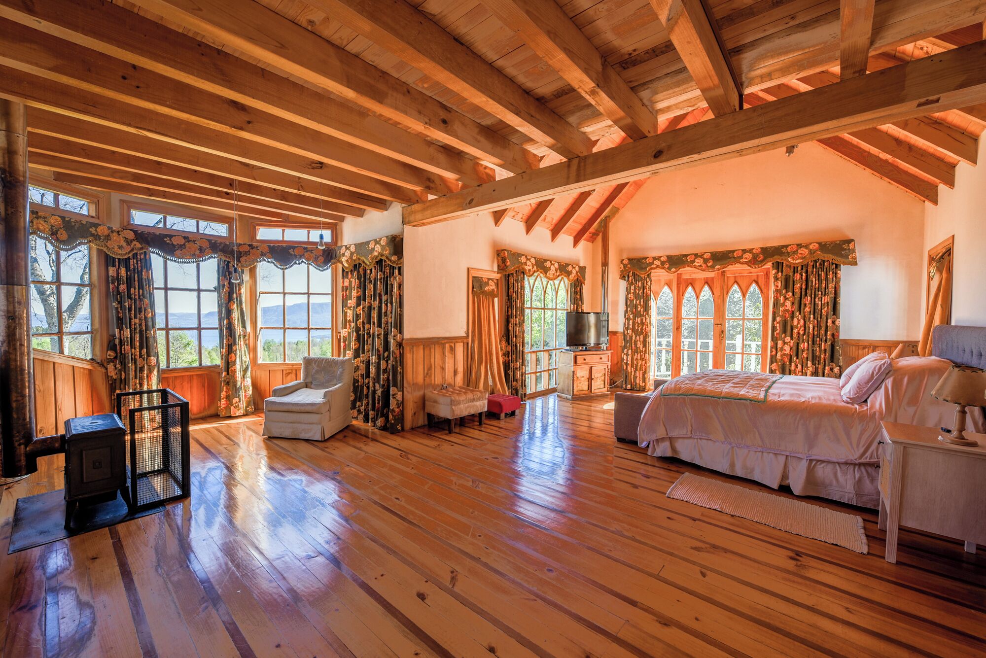 Une pièce spacieuse avec un plancher en bois, un lit, une télévision et de grandes fenêtres dotées de rideaux.