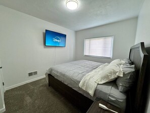 A bedroom with a bed, a television mounted on the wall, a window with blinds, and a carpeted floor.
