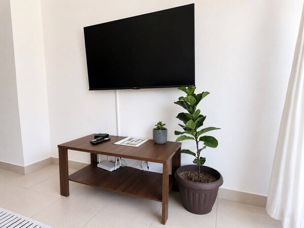 A room with a flat-screen TV mounted on the wall, a small wooden table with a plant, and a potted plant beside the table.