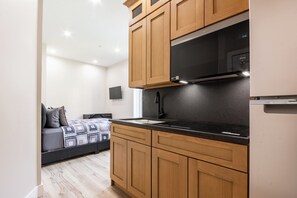 A compact kitchen with wooden cabinets and a black countertop, a microwave above the stove, and a bed with patterned bedding in the background. - Charming 1-bedroom apartment with AC in vibrant Brooklyn (Brooklyn)