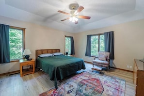 Quarto com cama, cadeira, mesa de cabeceira, abajur, ventilador de teto e janela.