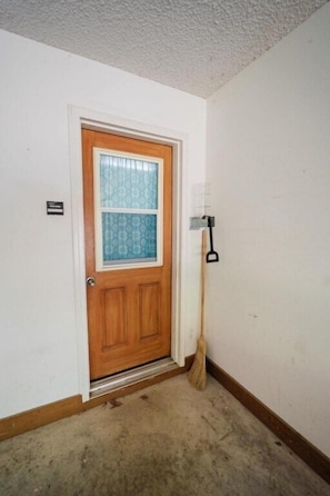 Uma porta de madeira fechada com uma janela, uma vassoura encostada na parede e um interruptor de luz na parede.