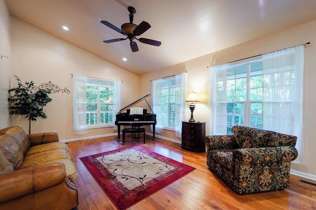 A spacious living room with a grand piano, a ceiling fan, and a large window with a view of trees.