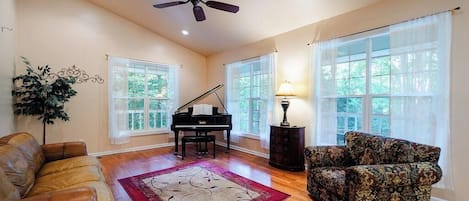 A spacious living room with a grand piano, a ceiling fan, and a large window with a view of trees.