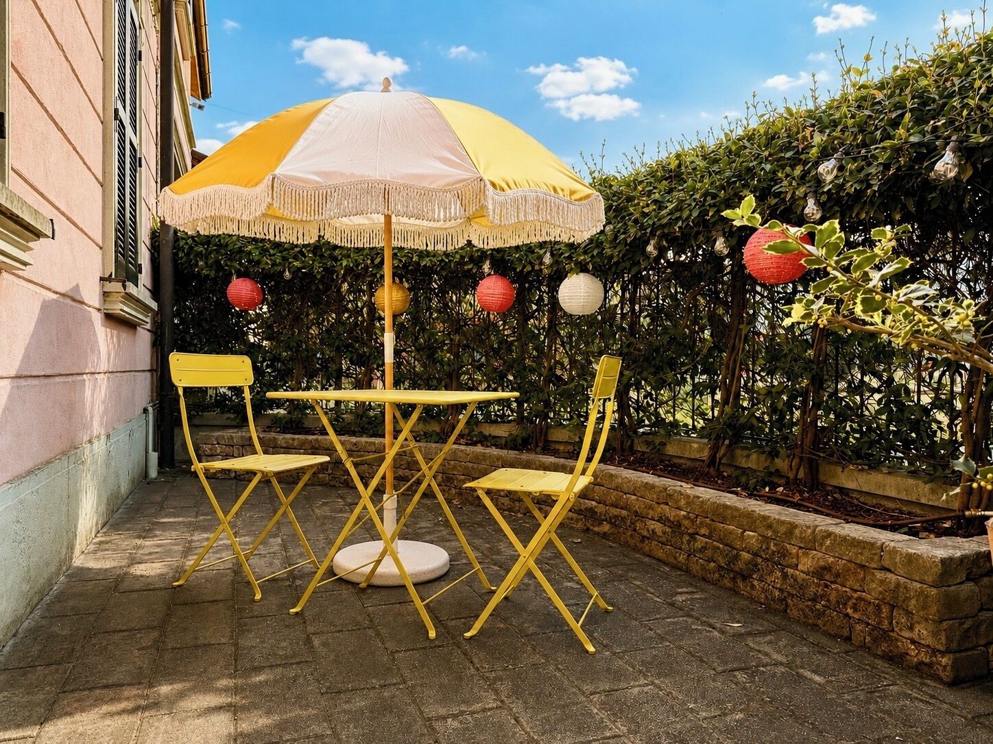 Een terras buiten met een gele tafel en stoelen, een wit en geel parasol en decoratieve lantaarns.