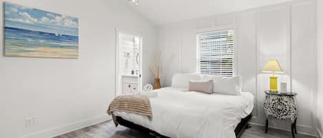 Un dormitorio con cama, ventilador de techo, ventana con persianas, mesita de noche con lámpara y un cuadro en la pared.