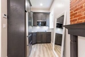 A modern kitchen with dark cabinetry, stainless steel appliances, and a brick accent wall. - Elegant Studio with Fireplace & Modern Comforts in Brooklyn (Brooklyn)