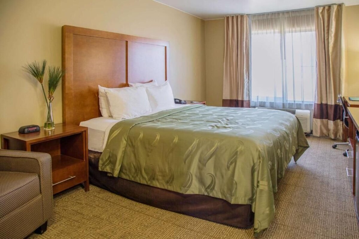 Habitación de hotel con una cama grande, una mesita de noche, una silla, un escritorio y un ventanal con cortinas.