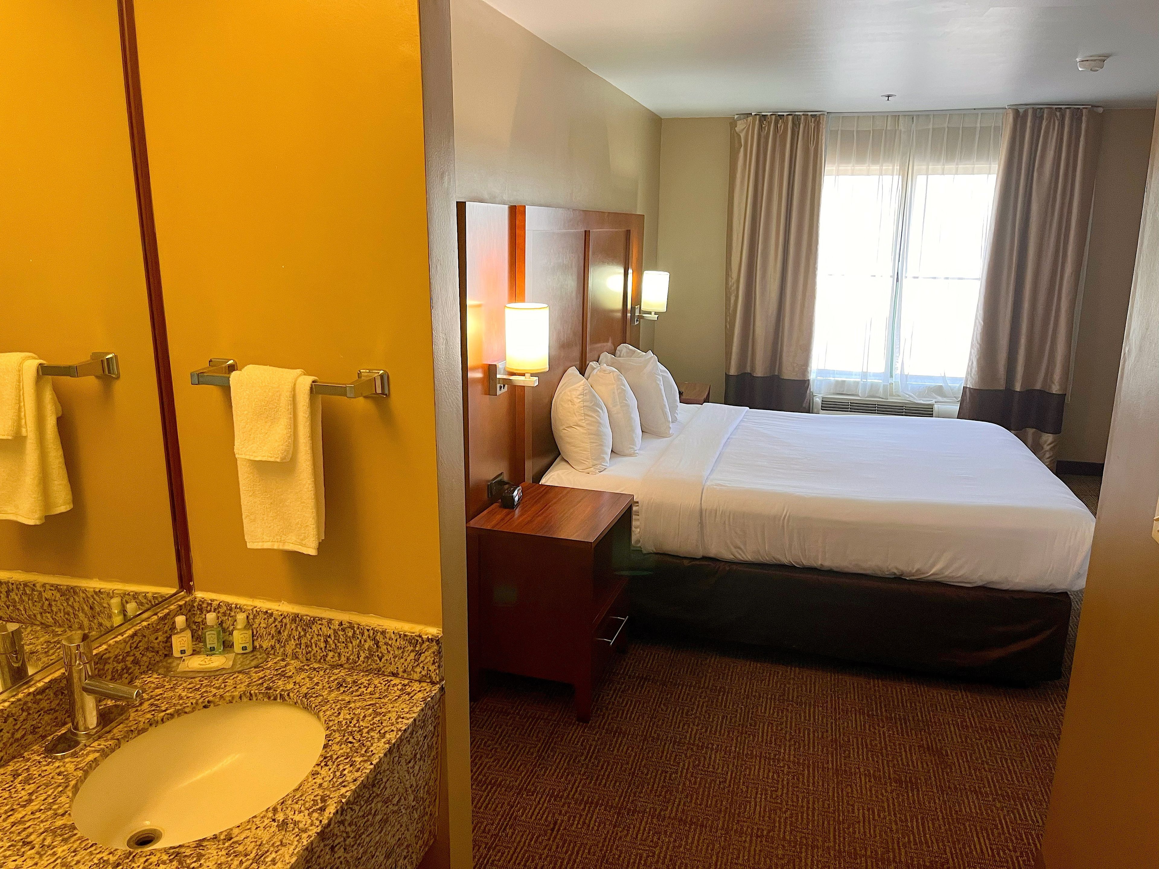 Une chambre d’hôtel avec un lit, une salle de bain comprenant un lavabo et un miroir, et une fenêtre avec des rideaux.