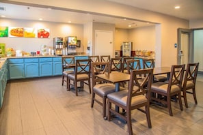 Un comedor de hotel con mesas y sillas de madera, una barra de cocina con diversos alimentos y un refrigerador.