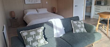 Quarto moderno com cama, sofá verde com almofadas de palmeira, manta branca e área de cozinha com micro-ondas.