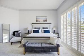 Ein Schlafzimmer mit einem großen Bett, einer Bank, einem Sessel, einem Nachttisch, einer Lampe, einem Spiegel und einem Fenster mit Jalousien.