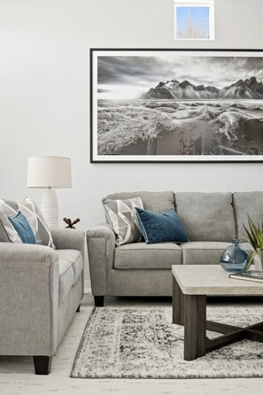 Ein Wohnzimmer mit grauem Sofa, einem Couchtisch, einer Lampe und einem gerahmten Landschaftsbild an der Wand.