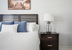 Un lit bien fait, avec une tête de lit, une table de chevet avec une lampe et un tableau encadré au-dessus du lit.