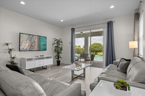 A modern living room with a sofa, coffee table, TV, and a view of a pool. - Encore 10BR Escape with Pool, Spa & Game Room (Kissimmee)