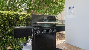 En udendørs grill med kontrolpanel, tilsluttet gasbeholder, foran grøn vegetation og et skilt med regler for poolen.