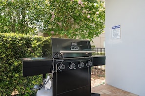 An outdoor grill with a control panel, connected to a gas tank, set against a backdrop of greenery and a pool rules sign. - Encore 10BR Escape with Pool, Spa & Game Room (Kissimmee)