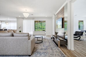 Ein modernes Wohnzimmer mit einem großen Sofa, einem Couchtisch und einem Fernseher. Ein großes Fenster bietet Blick auf viel Grün draußen.