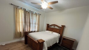 Ein Schlafzimmer mit einem Holzbett, einem Nachttisch, einer Fensterbank, einem Deckenventilator und einem Fenster mit Vorhängen.