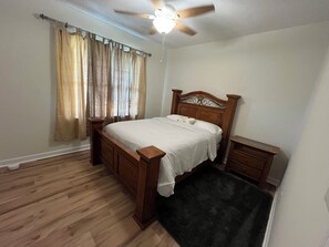 Ein Schlafzimmer mit einem Holzbett, einem Nachttisch, einer Fensterbank, einem Deckenventilator und einem Fenster mit Vorhängen.