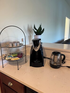 Eine Küchenarbeitsplatte mit Kaffeemaschine, einem Glas-Wasserkocher und einem Metallständer mit Teebeuteln und einer Topfpflanze.