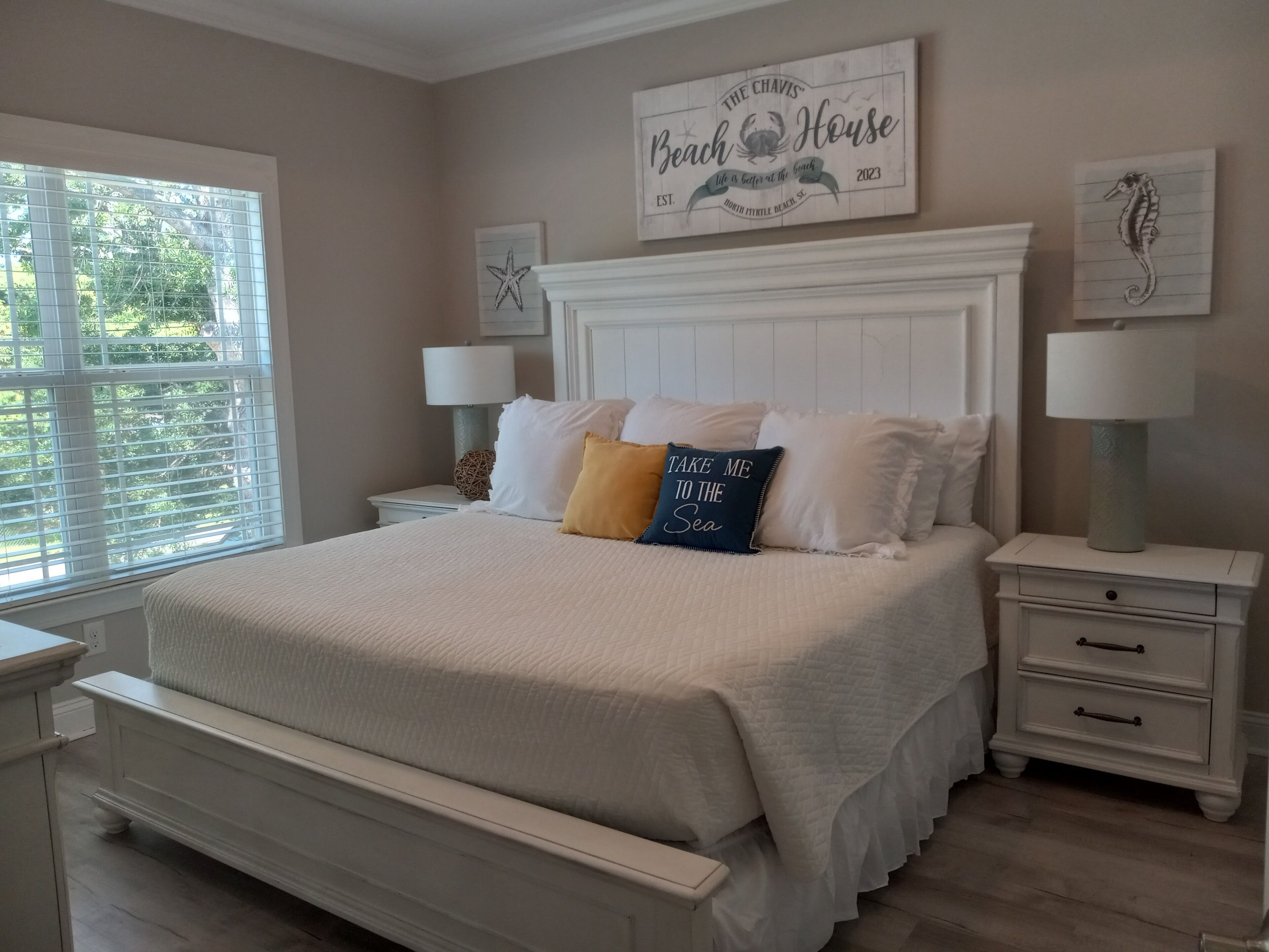 A bedroom with a large bed, white bedding, two bedside tables with lamps, a decorative sign above the bed, and a window with blinds.