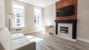 Een woonkamer met een bank, een open haard, een televisie aan een bakstenen muur en grote ramen.