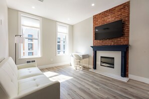 A living room with a sofa, a fireplace, a TV mounted on a brick wall, and large windows. - Stylish Brooklyn Apartment Natural Light (Brooklyn)