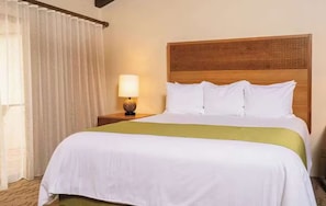 A hotel room with a bed, a nightstand, a lamp, and a window with curtains. - Spacious 3-bedroom condo in a peaceful location with AC and gym (Kailua-Kona)