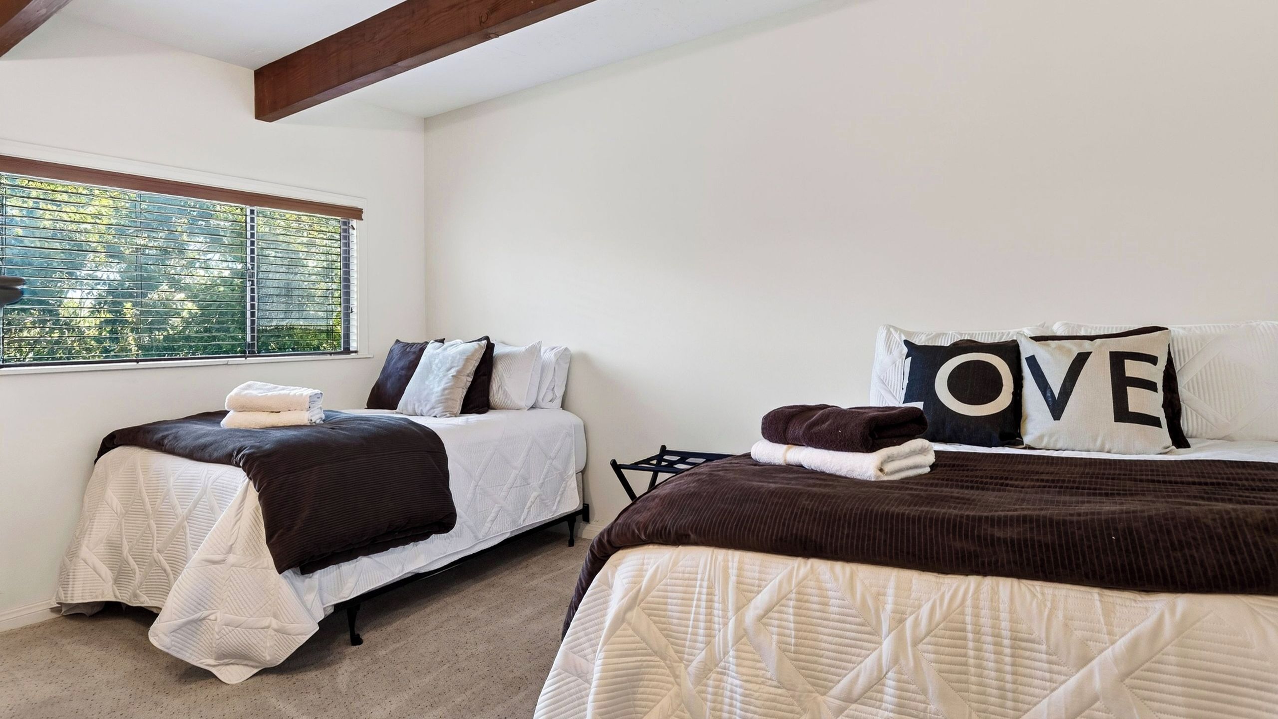 A bedroom with two beds, each with a quilted bedspread and pillows, a window with blinds, and a view of greenery outside.