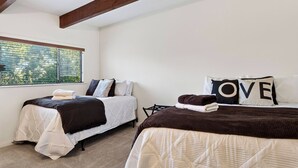 A bedroom with two beds, each with a quilted bedspread and pillows, a window with blinds, and a view of greenery outside. - Golden Hour Lodge by Avantstay BBQ + Great Room + Multiple Decks Lake & Sunset Views (Lake Arrowhead)