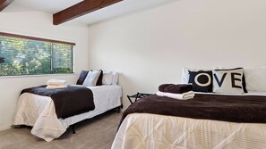 A bedroom with two beds, each with a quilted bedspread and pillows, a window with blinds, and a view of greenery outside. - Golden Hour Lodge by Avantstay BBQ + Great Room + Multiple Decks Lake & Sunset Views (Lake Arrowhead)