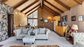 A cozy living room with a stone wall, a fireplace, a dining area, and a kitchen with wooden cabinets. - Golden Hour Lodge by Avantstay BBQ + Great Room + Multiple Decks Lake & Sunset Views (Lake Arrowhead)
