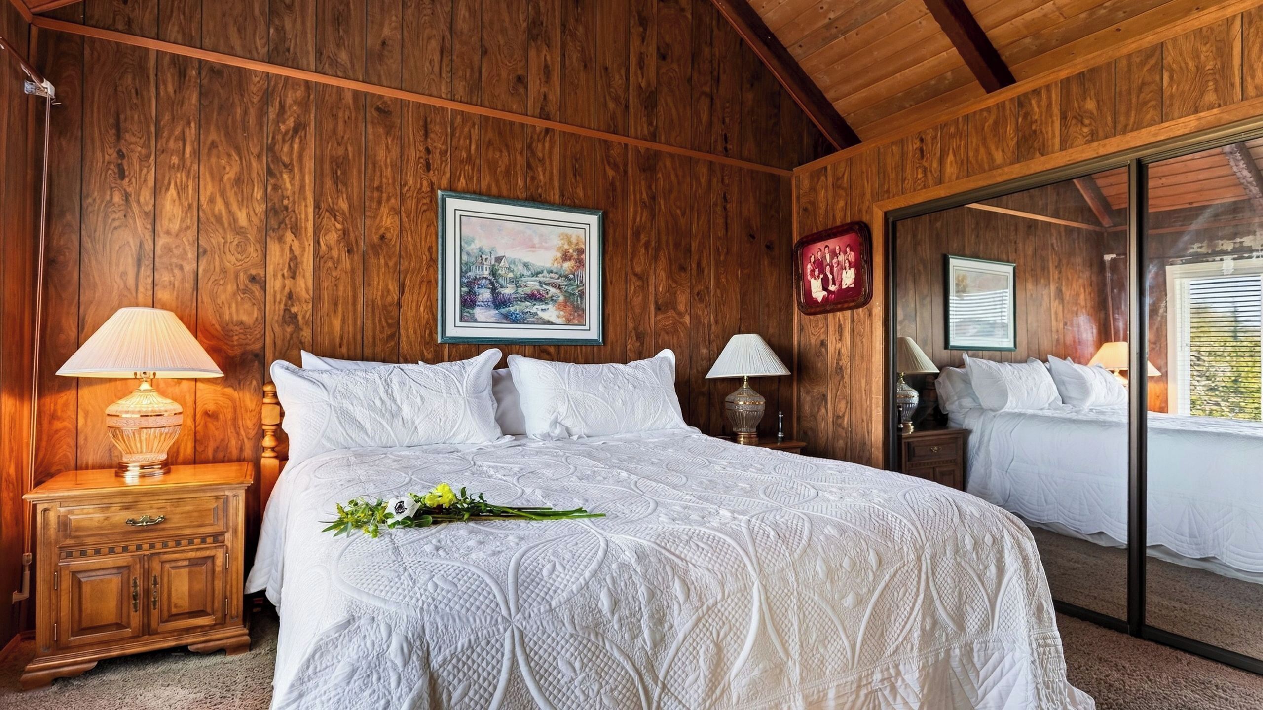 A bedroom with a large bed, wooden walls, a nightstand with a lamp, and a mirror reflecting the room.