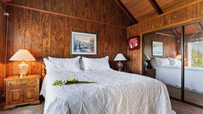 A bedroom with a large bed, wooden walls, a nightstand with a lamp, and a mirror reflecting the room.