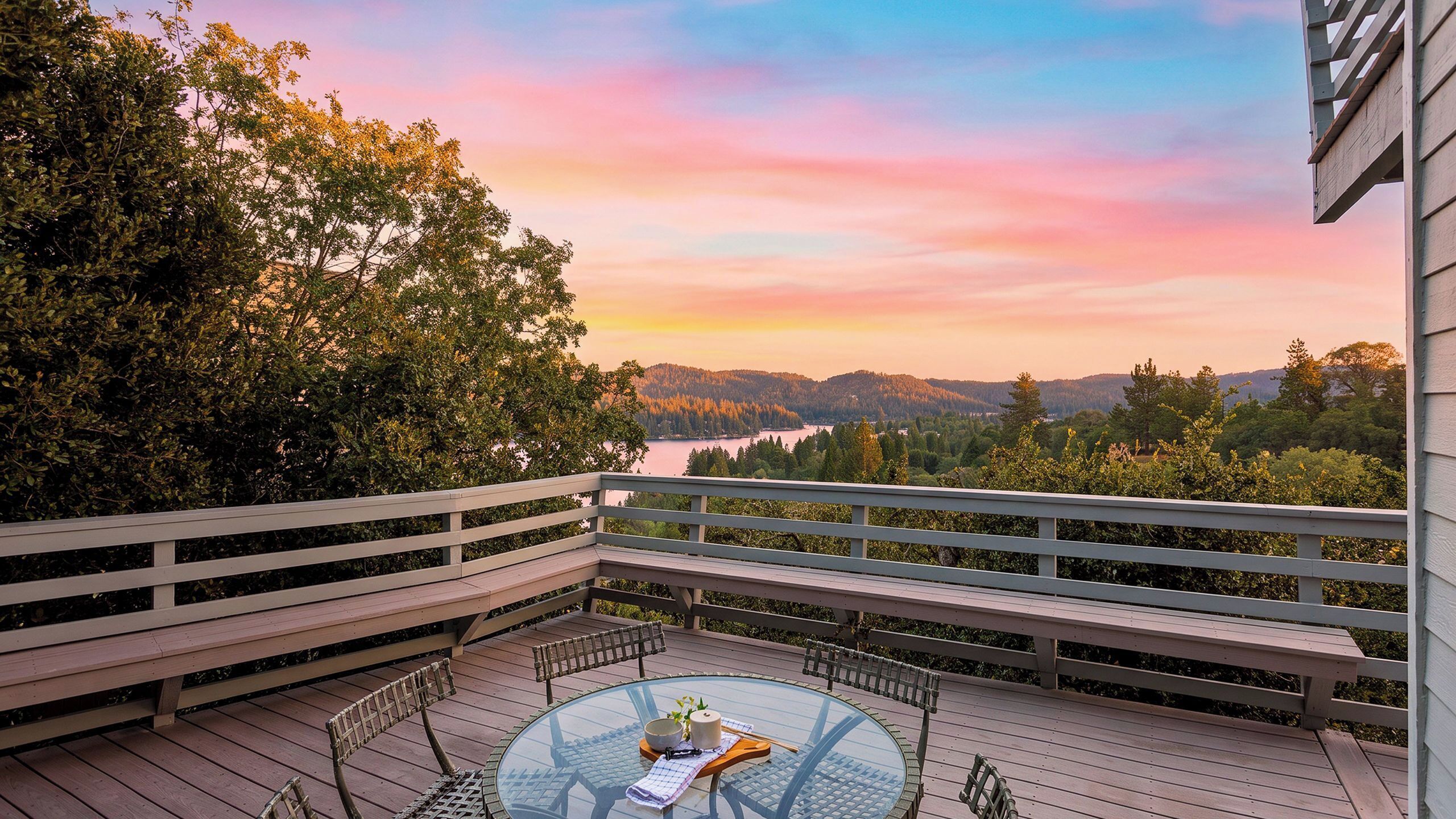 Une terrasse dotée d’une table à plateau en verre, de bancs et offrant une vue sur un lac et une forêt au coucher du soleil.