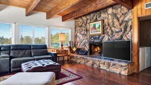 A living room with a stone fireplace, a flat-screen TV, and a leather sofa. - Golden Hour Lodge by Avantstay BBQ + Great Room + Multiple Decks Lake & Sunset Views (Lake Arrowhead)