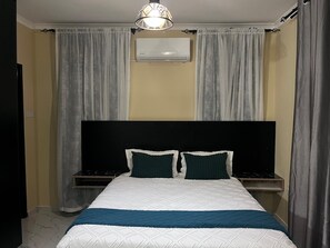 A bedroom with a bed, two bedside tables, a wall-mounted air conditioner, and a ceiling light fixture.