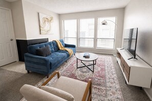 A living room with a blue sofa, a small round table, a television on a stand, and a patterned rug. - Sleepover | Prime 1BD/1BA + Gym - Wichita (Wichita)