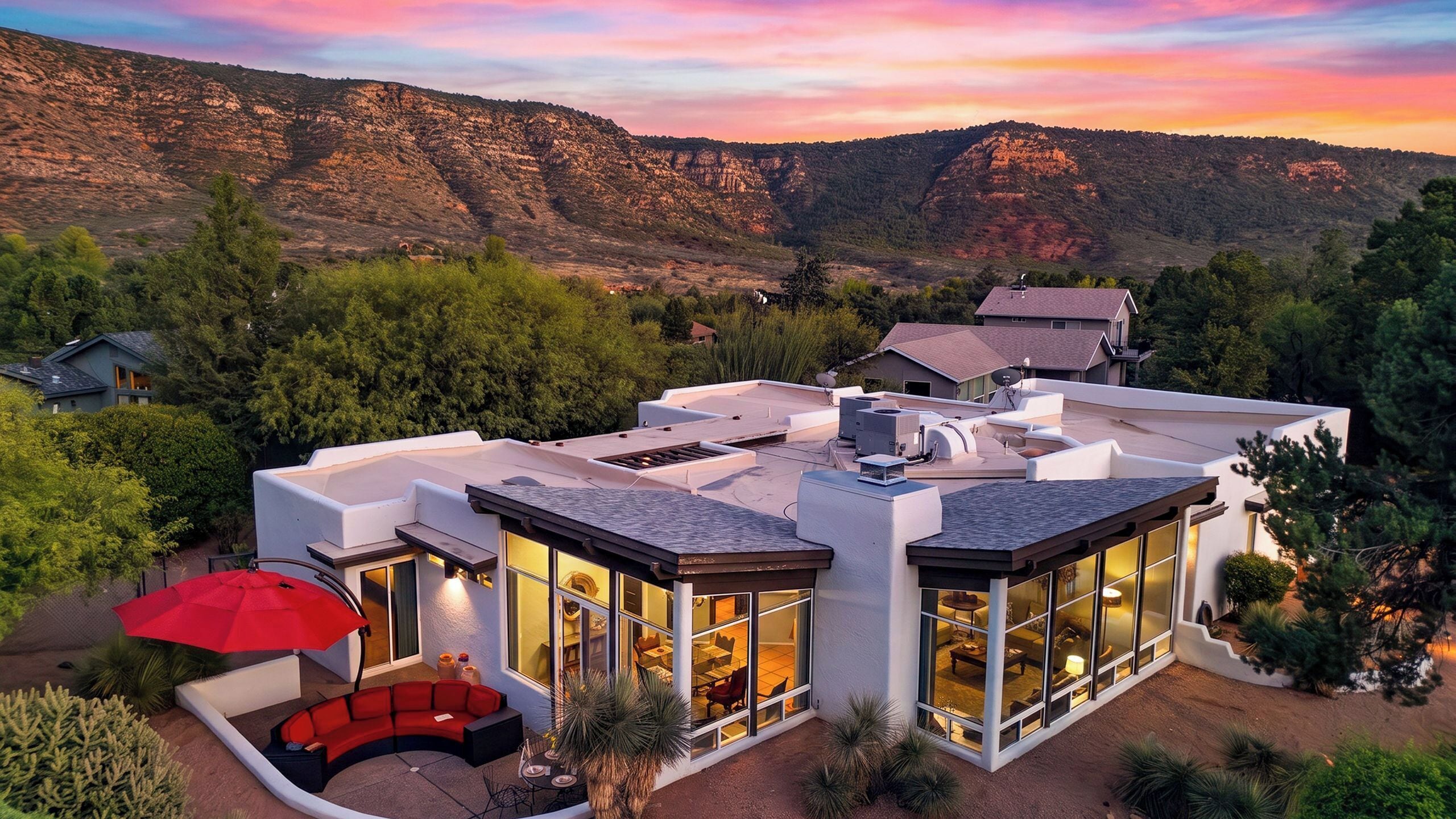 Una casa moderna con una amplia fachada de vidrio, un patio cubierto con muebles rojos y vistas a las montañas circundantes.
