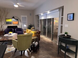 Ein Wohnzimmer mit einer Couch, Sesseln, einem Couchtisch und einem Fernseher. Eine Glastür führt auf eine Terrasse.
