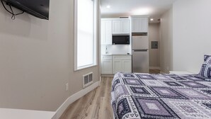 Ein Schlafzimmer mit einem Bett, einem an der Wand montierten Fernseher, einem Fenster und einer Küchenzeile im Hintergrund.
