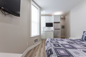 Ein Schlafzimmer mit einem Bett, einem an der Wand montierten Fernseher, einem Fenster und einer Küchenzeile im Hintergrund.