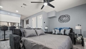 A modern bedroom with a large bed, a ceiling fan, and a dining area with bar stools. - Walk to AT&T Stadium | Modern 2BR, Free Parking (Arlington)