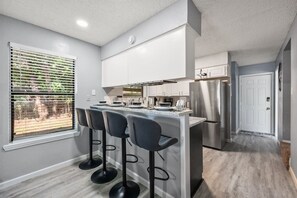 A modern kitchen with a central island, bar stools, stainless steel appliances, and a window with blinds. - Walk to AT&T Stadium | Modern 2BR, Free Parking (Arlington)