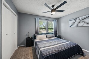 A bedroom with a bed, a nightstand, a ceiling fan, a window with curtains, and a wall-mounted abstract art piece. - Walk to AT&T Stadium | Modern 2BR, Free Parking (Arlington)